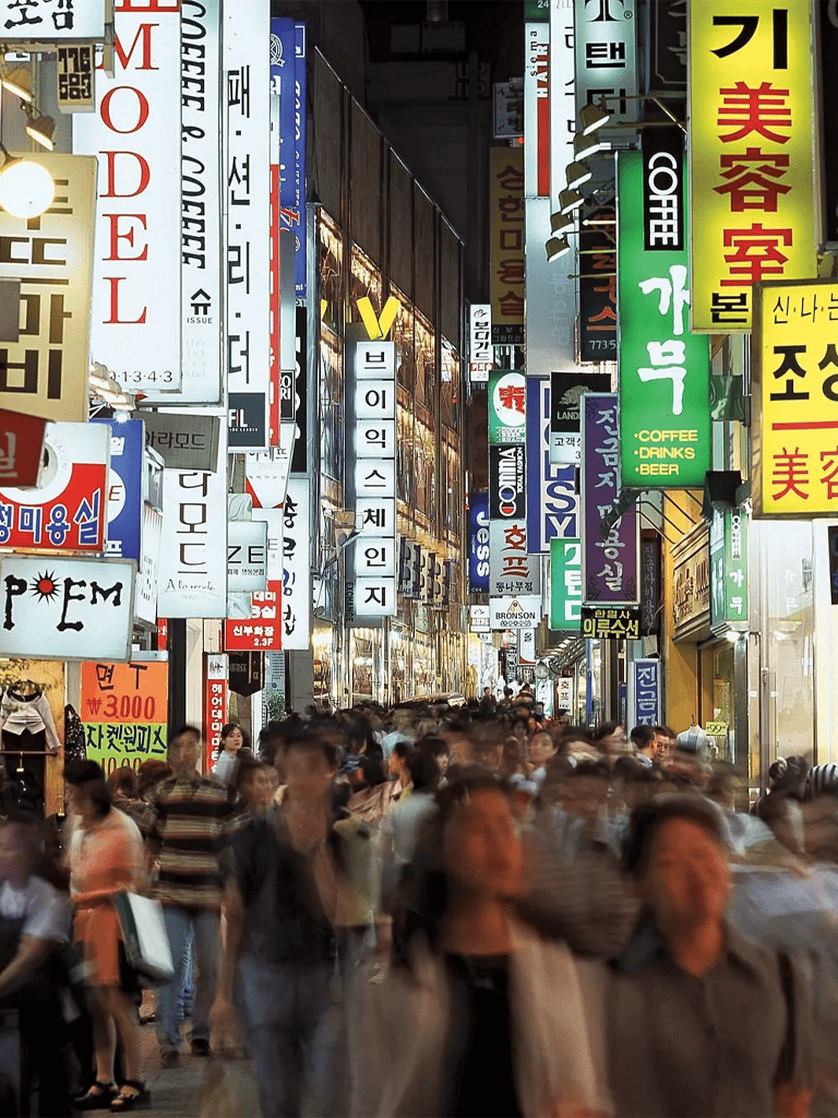 Bustling night street filled with neon signs advertising cafes, beauty salons, and various shops in Seoul, South Korea.