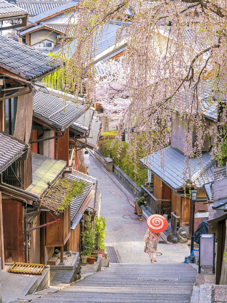 Cherry blossom street in Japan with traditional wooden houses and women with umbrellas, scenic springtime atmosphere.