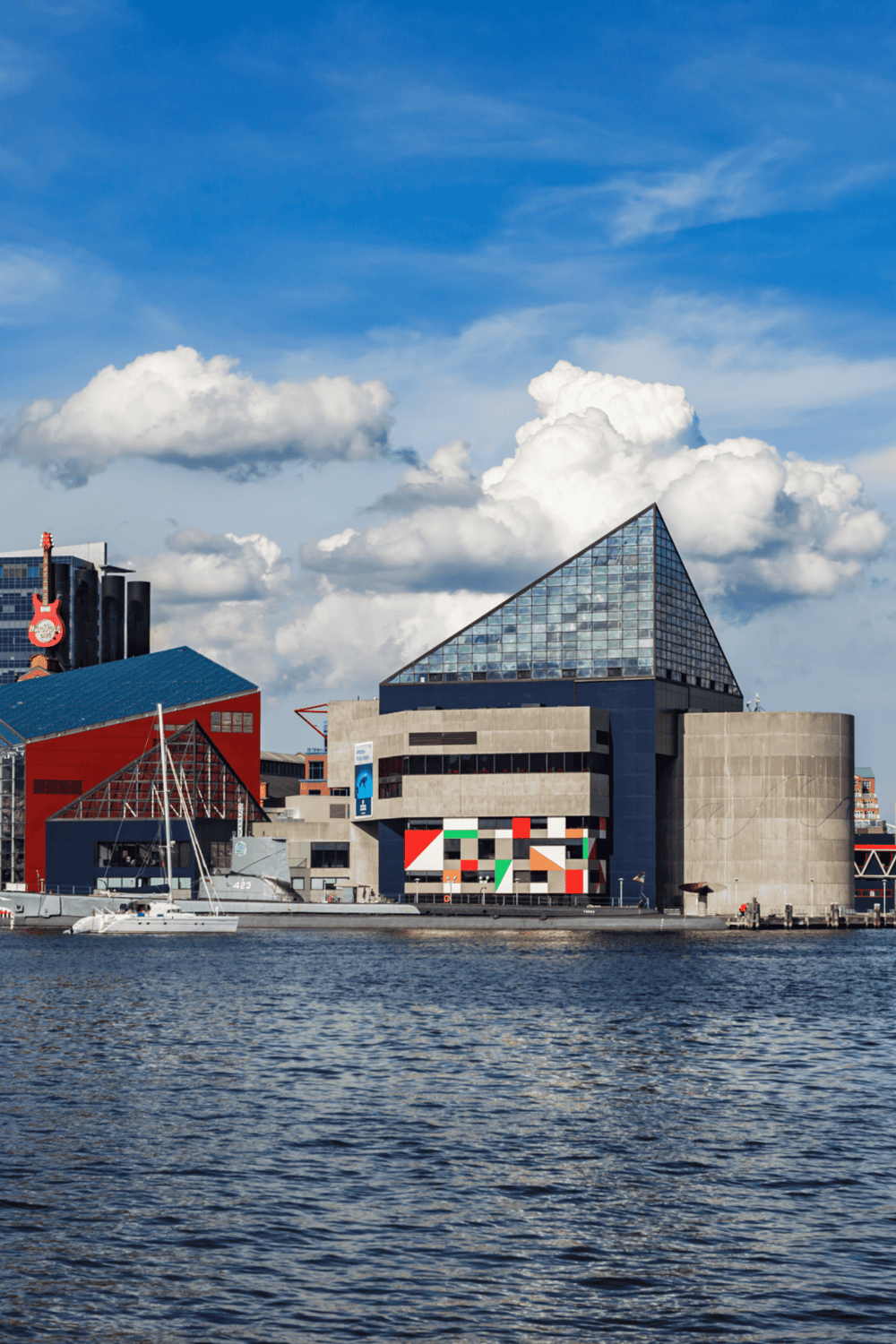 Modern urban waterfront with unique architectural buildings and sailboats under a partly cloudy sky.