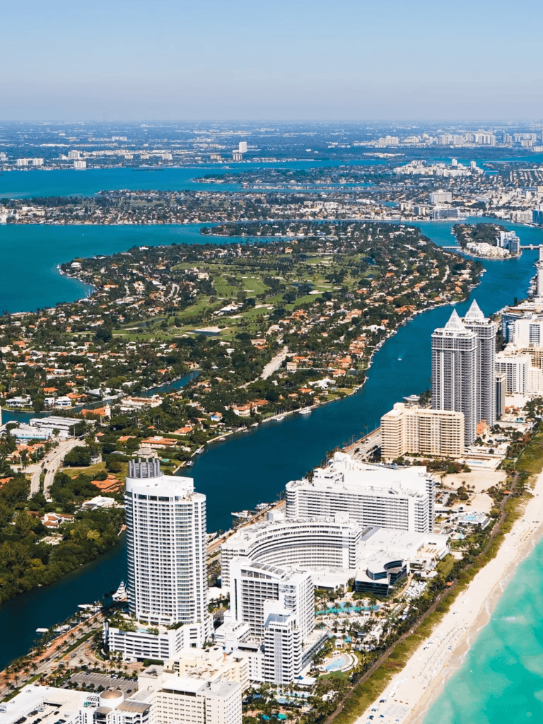 Aerial view of Miami beachfront cityscape and waterways with tall hotels and lush greenery.