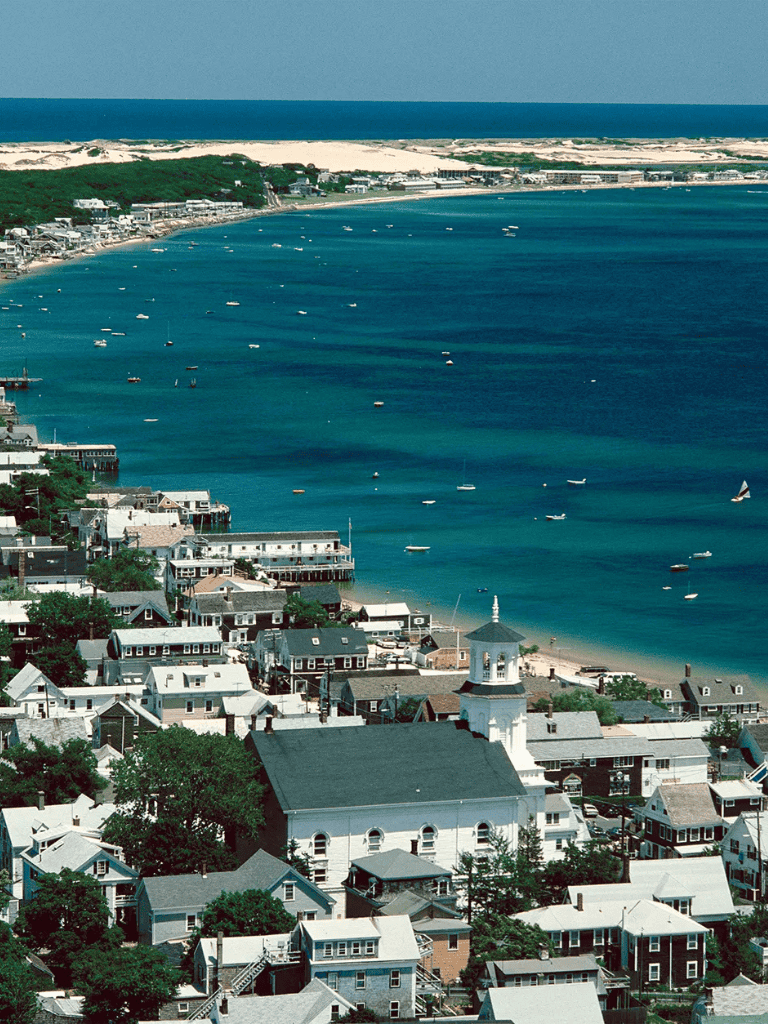 Beachfront residential houses and boats docked in a coastal town with dunes in the background.