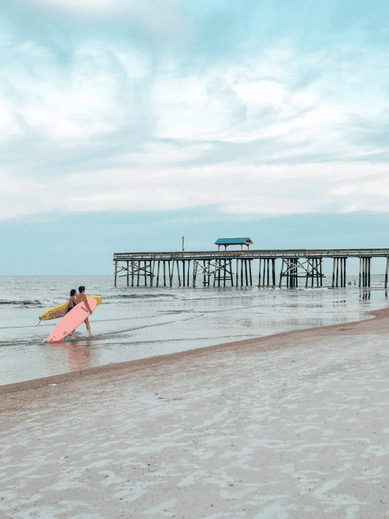 People carrying surfboards near the beach at a wooden pier under a cloudy sky.