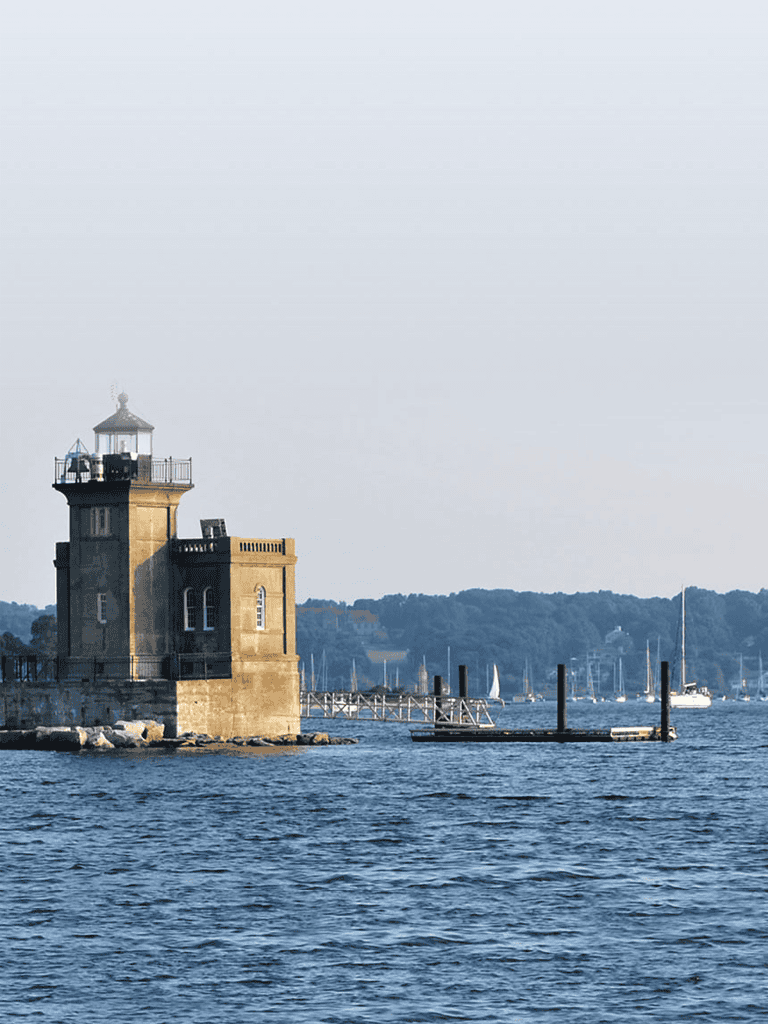 Lighthouse by the water with boats and a distant shoreline.