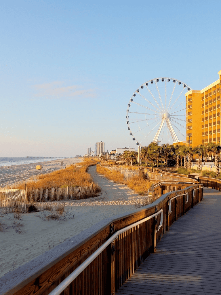 Seafront boardwalk with Ferris wheel and hotel buildings, clear blue sky, sunny coastline scenery.
