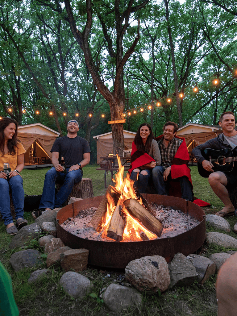 Cozy outdoor campfire gathering with friends at twilight, surrounded by tents and string lights.