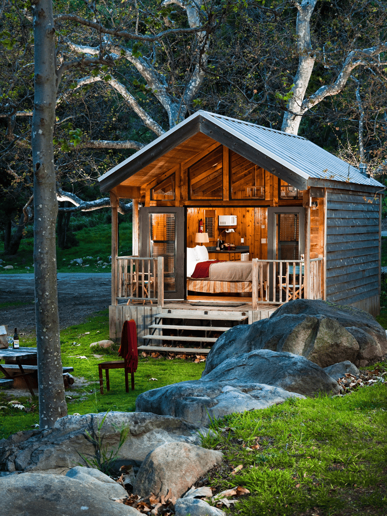 Cozy wooden cabin in forest with large rocks and outdoor seating, ideal for nature retreat.