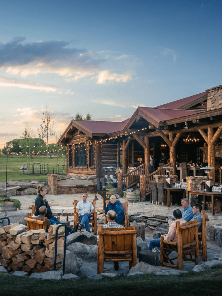 Outdoor rustic event venue with log cabin architecture and string lights, hosting a group discussion around a fire pit.