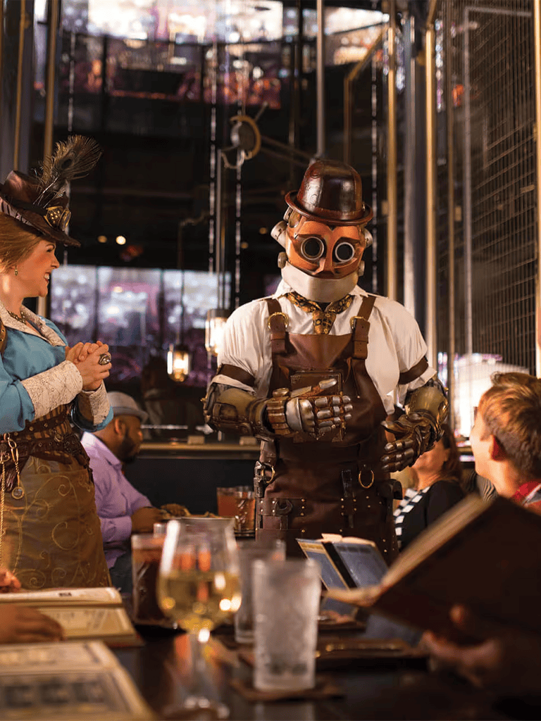 A steampunk-themed waiter with goggles and Victorian attire serving diners in a stylish restaurant.