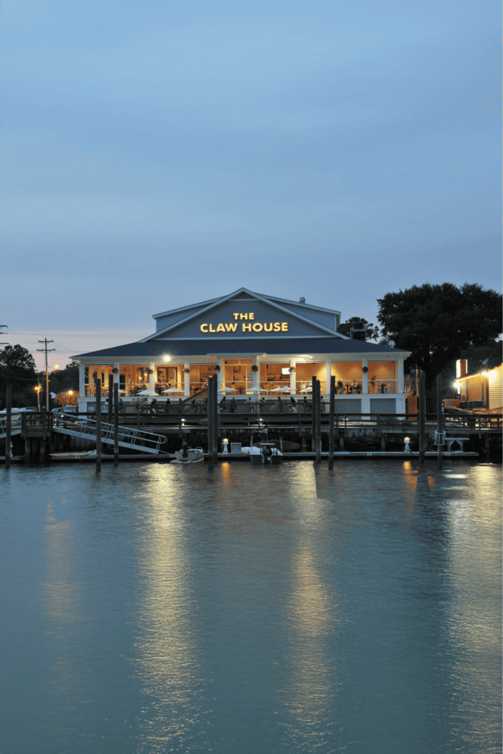 Quaint waterfront restaurant at dusk with illuminated "The Claw House" sign, dock, and boats in water.