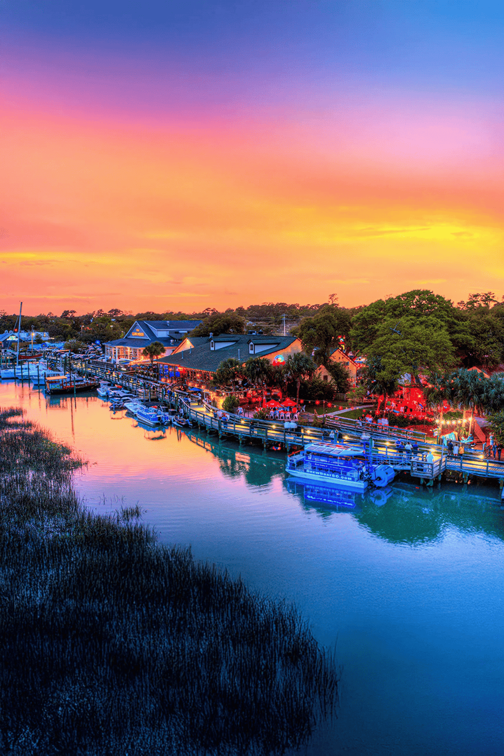 Vibrant marina at sunset with boats, waterfront dining, lush greenery, and colorful sky.