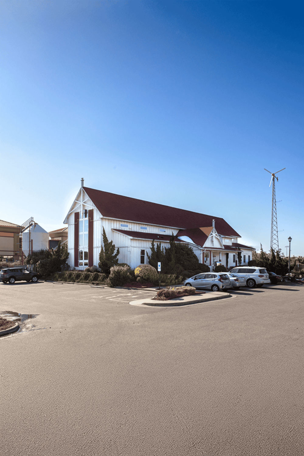 White church building with parking lot and wind turbine under clear blue sky.