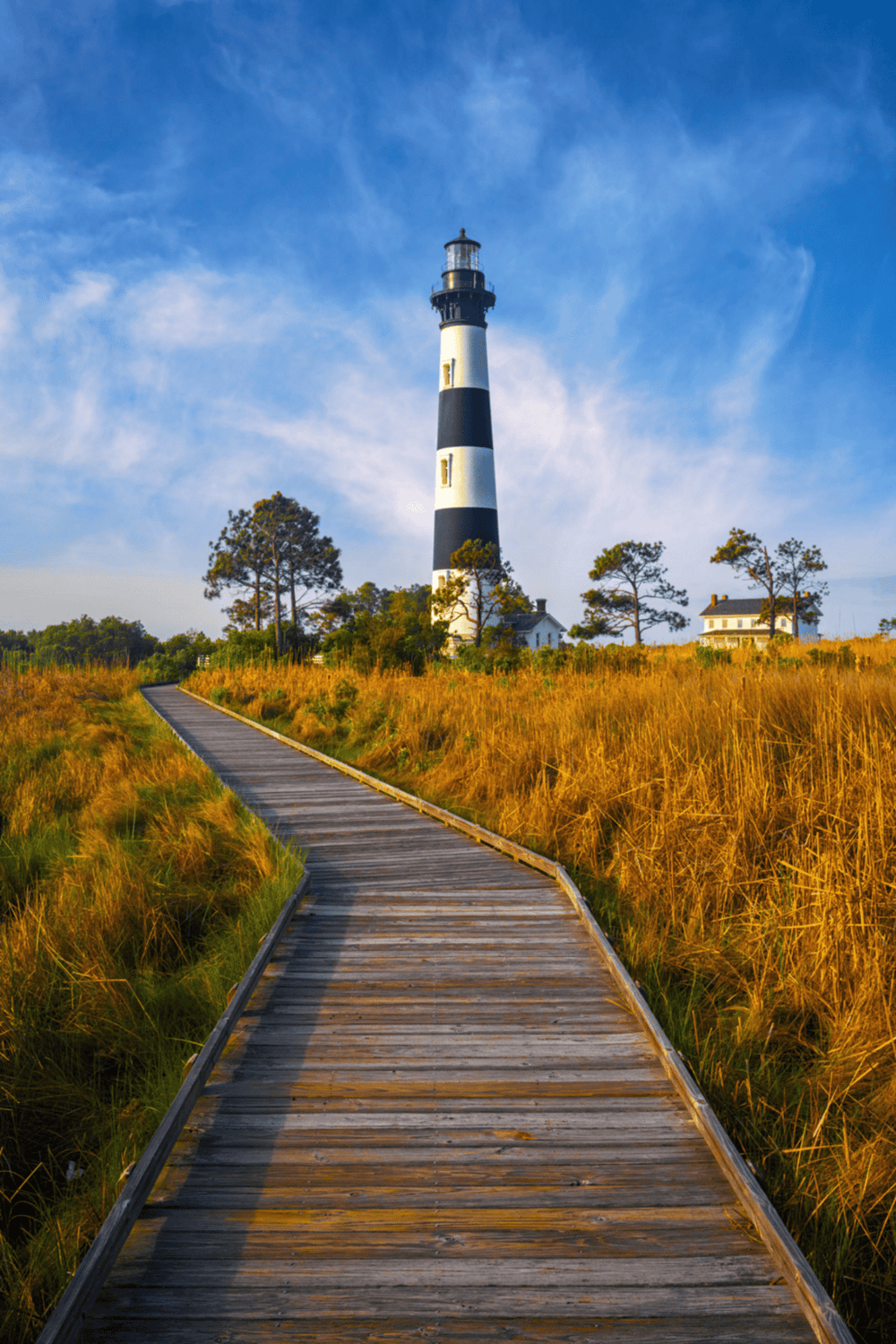 Lighthouse on coastal landscape with wooden boardwalk and golden grasses, scenic ocean view.