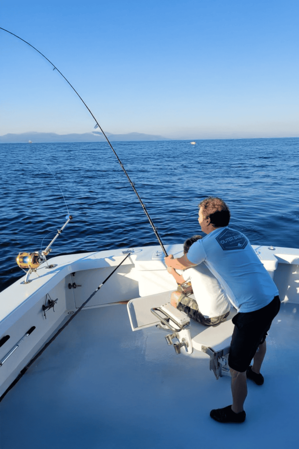 Gentlemen fishing on a boat in the ocean, clear blue sky, distant mountains, enjoying outdoor recreational activities.