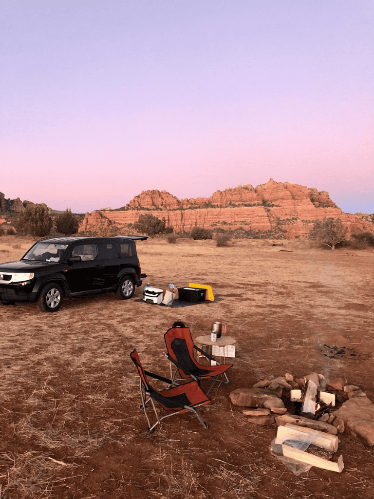 Desert campsite with chairs and camping gear at sunset with rocky formations in the background.