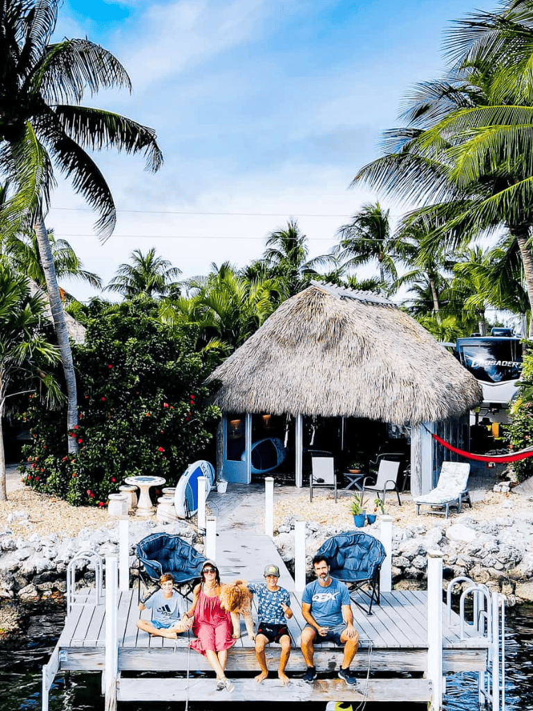 Relaxing tropical waterfront with palm trees, thatched hut, and friends sitting on dock by water.