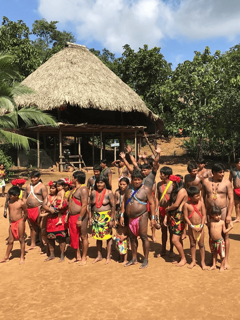Vibrant indigenous community gathered outdoors in traditional attire in a rural setting with thatched-roof hut.