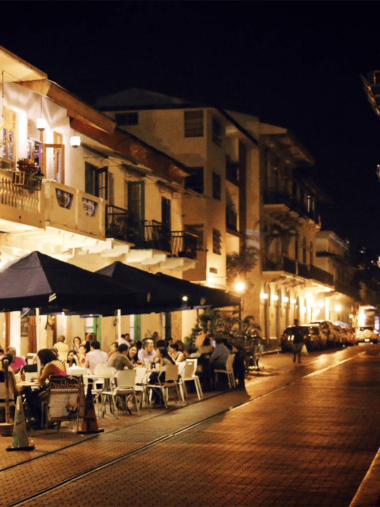 Nighttime street scene with outdoor dining, warm lights, and lively atmosphere in a charming urban area.