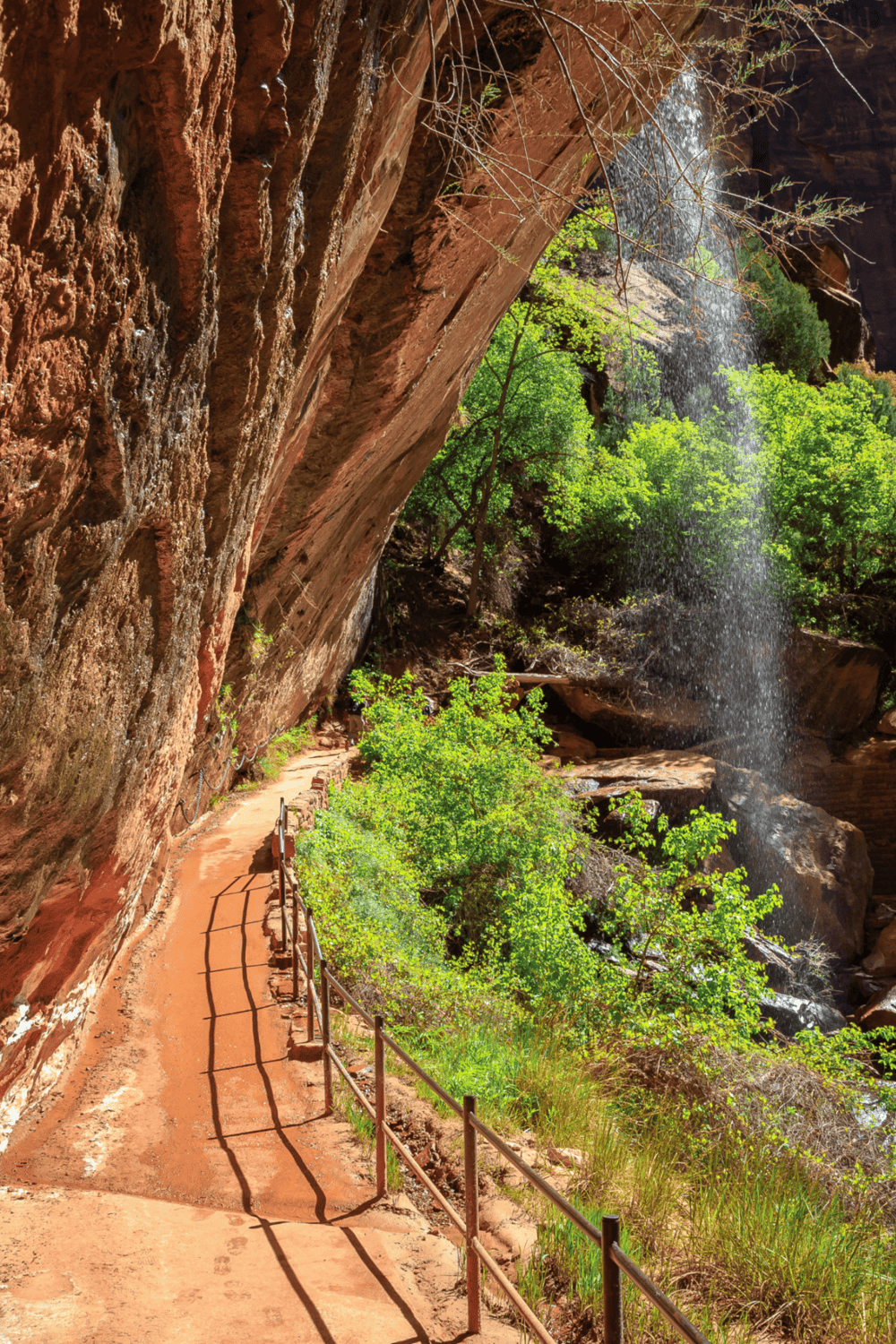 Vivid hiking trail along red rock cliffs with waterfall in a lush green canyon setting.