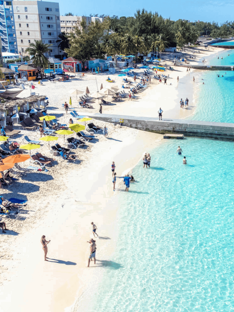 Sunny beach with umbrellas, lounging chairs, and people swimming and relaxing by the water.