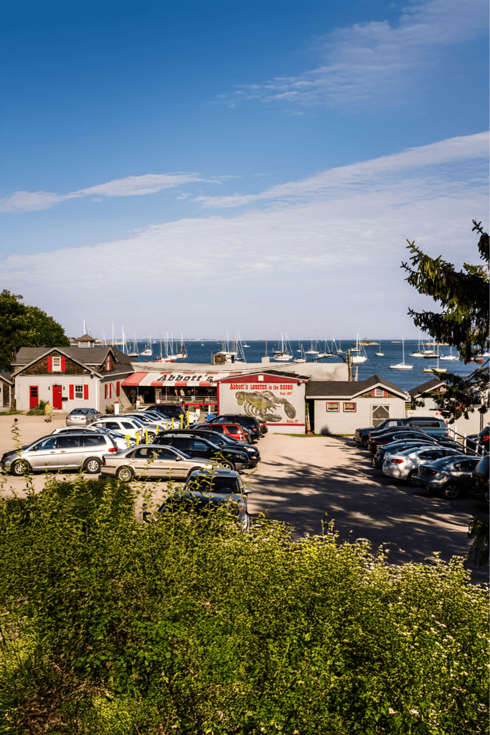 Coastal restaurant and marina with sailboats in the harbor, parking lot, and scenic ocean view.