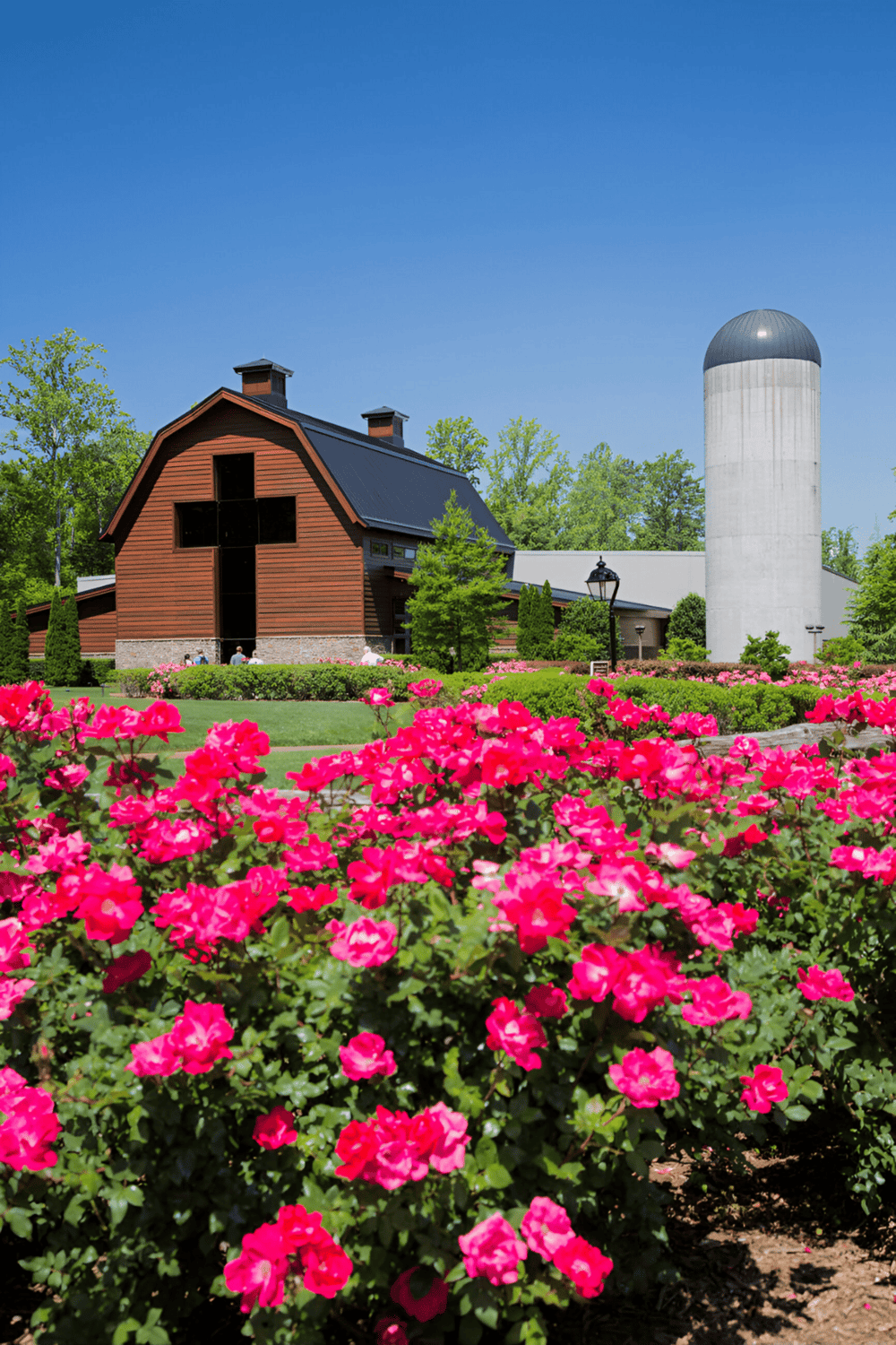 Rustic wooden barn with a cross-shaped window and a silo surrounded by colorful flowers and greenery.