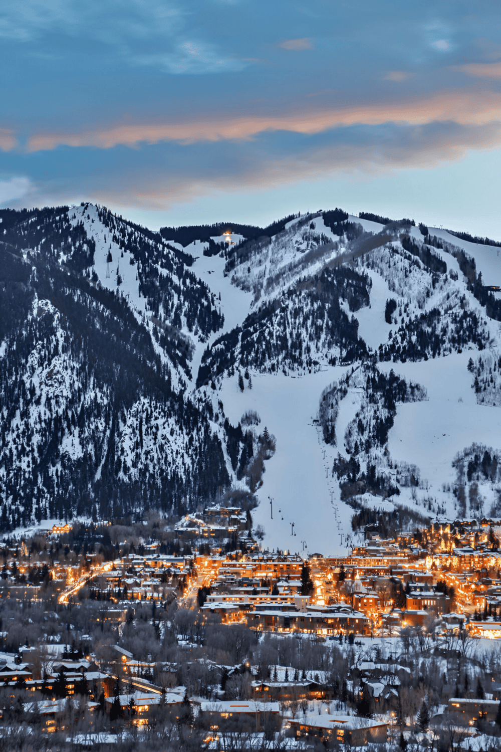 Snowy mountain resort with ski slopes and illuminated town at dusk.