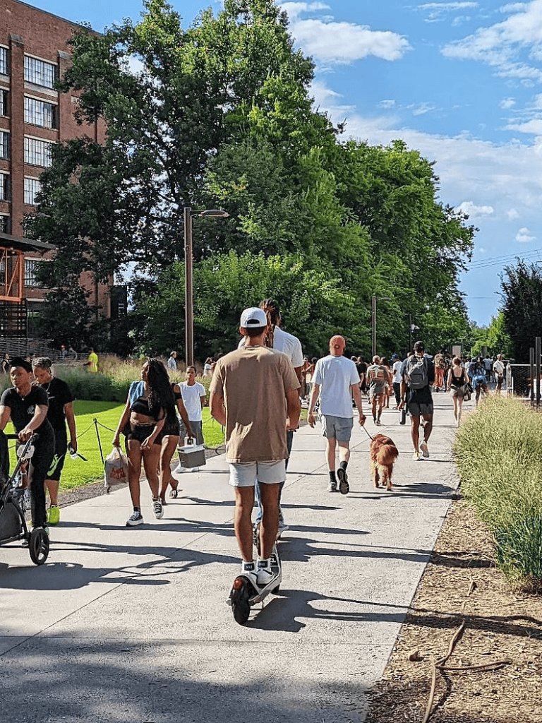 E-biker riding on city park pathway with pedestrians and greenery on sunny day.