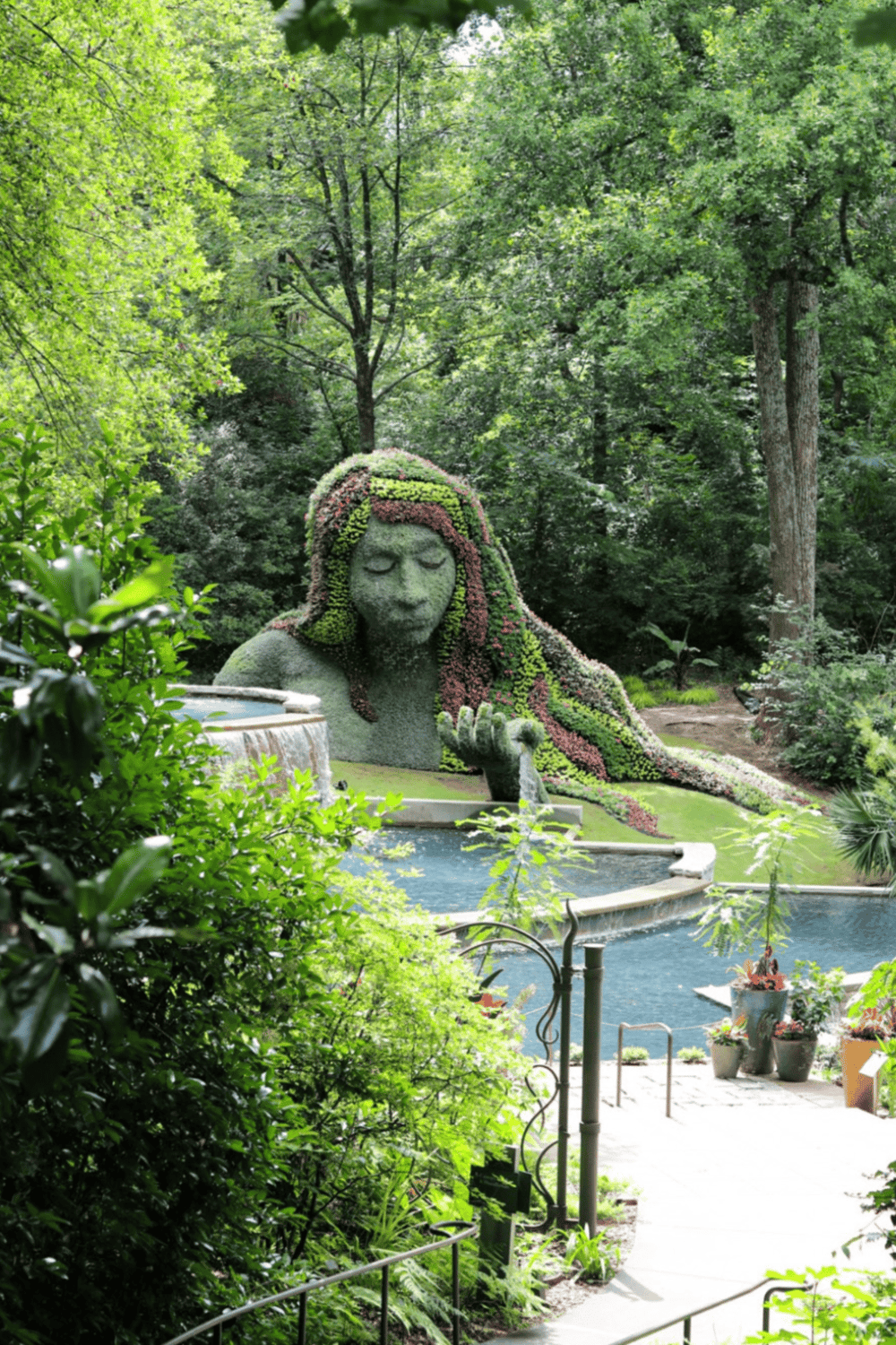 Lush green garden with a topiary of a woman’s face and flowing hair, surrounded by trees and flowers.