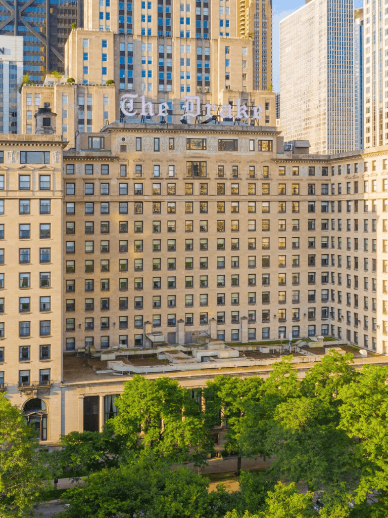 The Drake hotel in New York City with tall skyscrapers in the background and lush trees in the foreground.