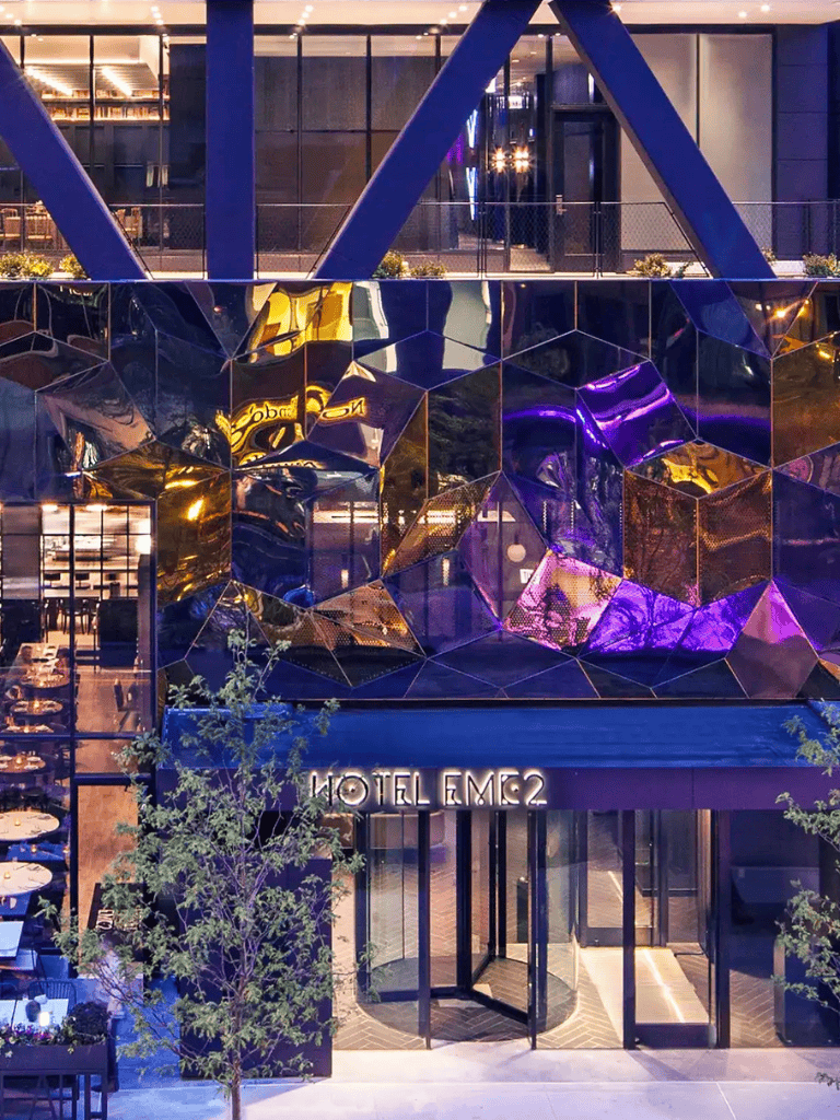 Modern hotel facade with reflective geometric art installation at night, illuminated by vibrant purple and yellow lights.