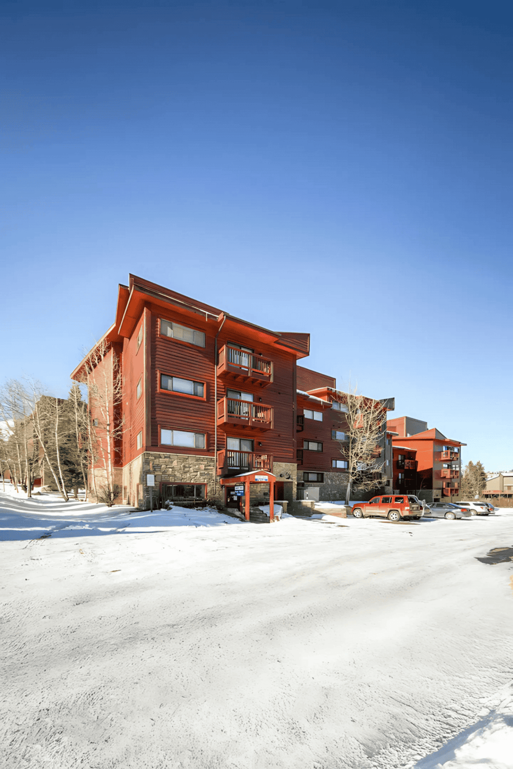 Modern red apartment building in snowy landscape with parked cars and leafless trees.