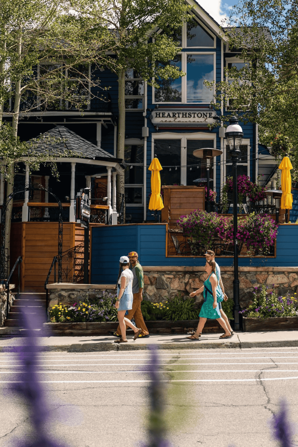 Blue house exterior with outdoor dining area and "Hearthstone" sign, vibrant flowers; scenic mountain town street.