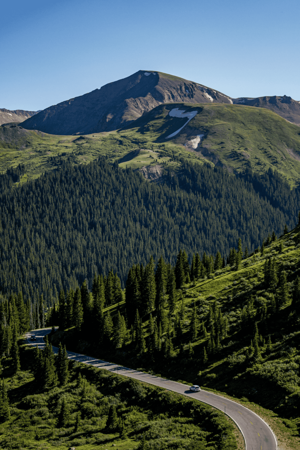 Serene mountain landscape with winding road, lush green forests, and clear blue sky.