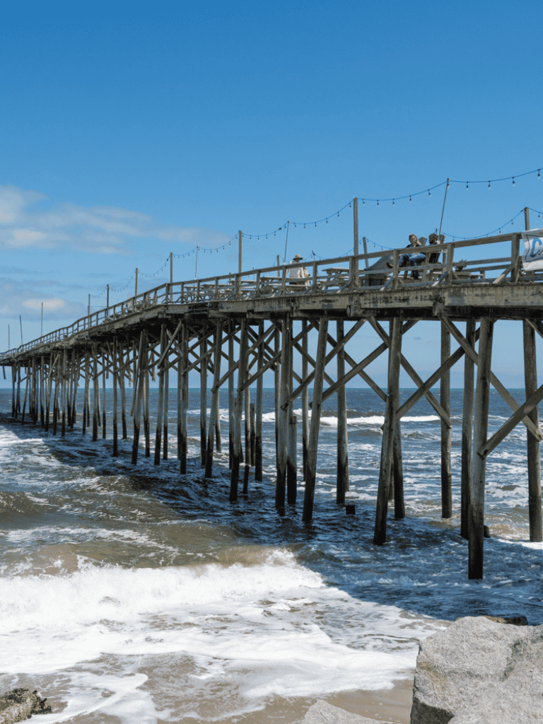 Weathered wooden pier over ocean with blue sky and string lights, embodying coastal leisure and scenic beauty.