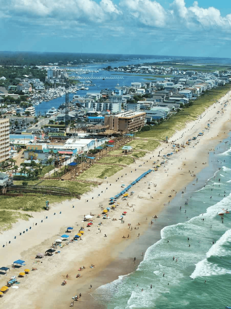 Aerial view of a coastal city featuring a sandy beach, ocean waves, and beachfront buildings with boats in the harbor.