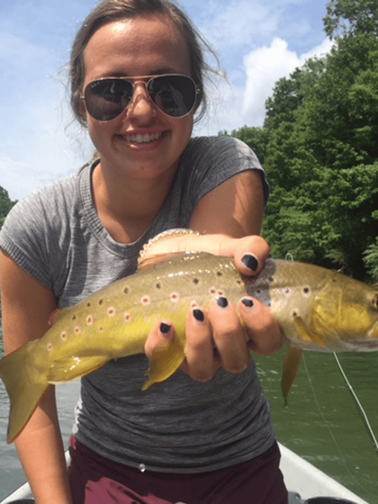 Large fish caught during outdoor fishing trip, woman smiling with sunglasses, lush green trees, sunny day, adventure.