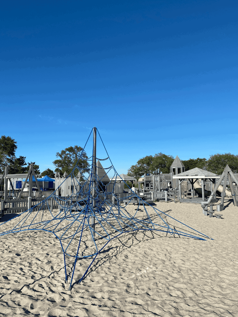 Bright blue sky over a well-designed children's playground with climbing structures and sandy ground.
