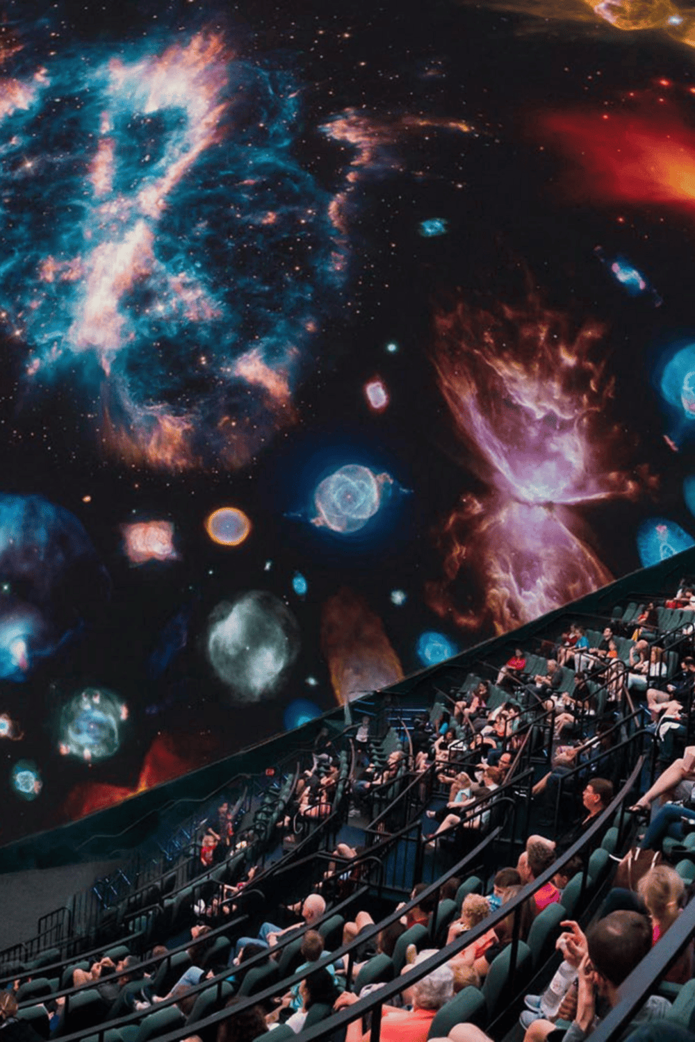 Vast galaxy-themed digital projection with audience watching a planetarium show or cinematic presentation.