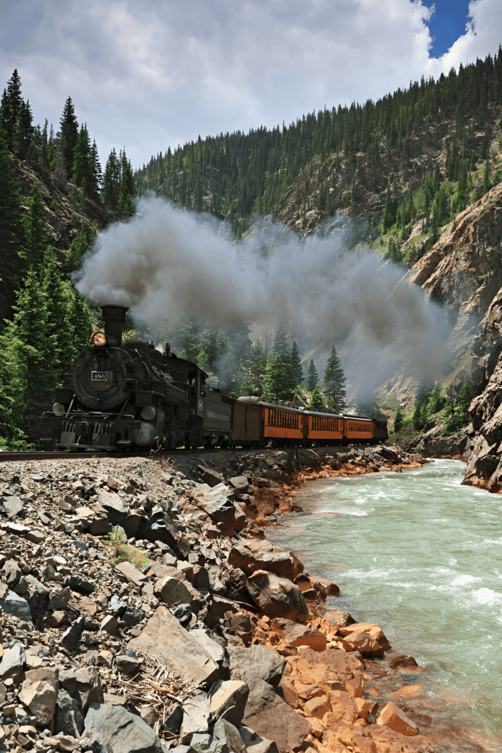 Steam locomotive train traveling through scenic mountain canyon with lush green trees and rocky shores.