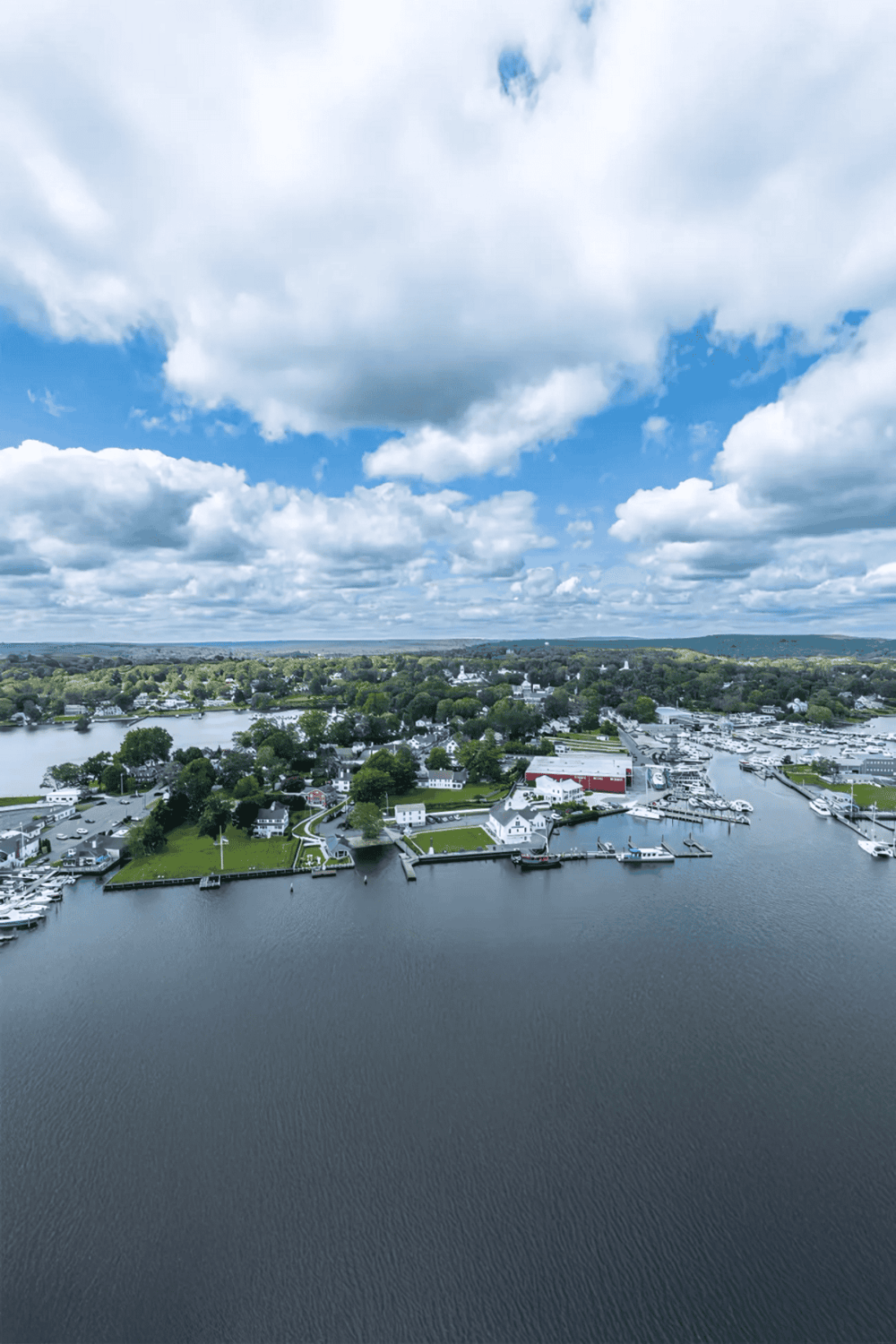 Aerial view of a scenic harbor town with boats and lush greenery under a partly cloudy sky.