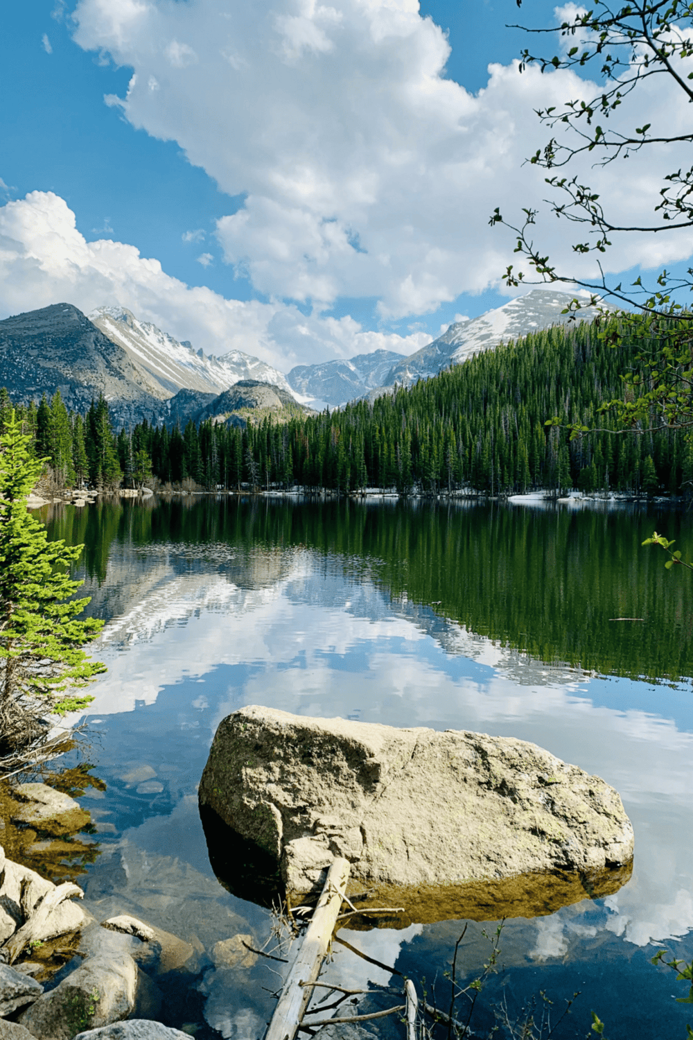 Serene mountain lake with lush pine trees, snow-capped peaks, and reflective water in a scenic wilderness setting.