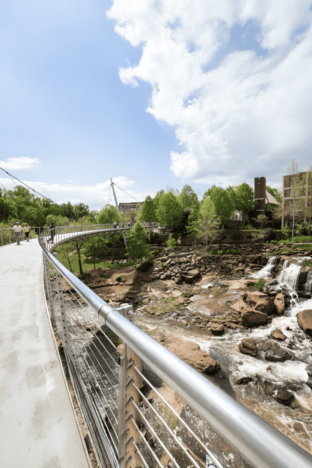 Scenic creekside park with pedestrian bridge, lush green trees, and waterfall. Perfect for outdoor exploration and nature walks.