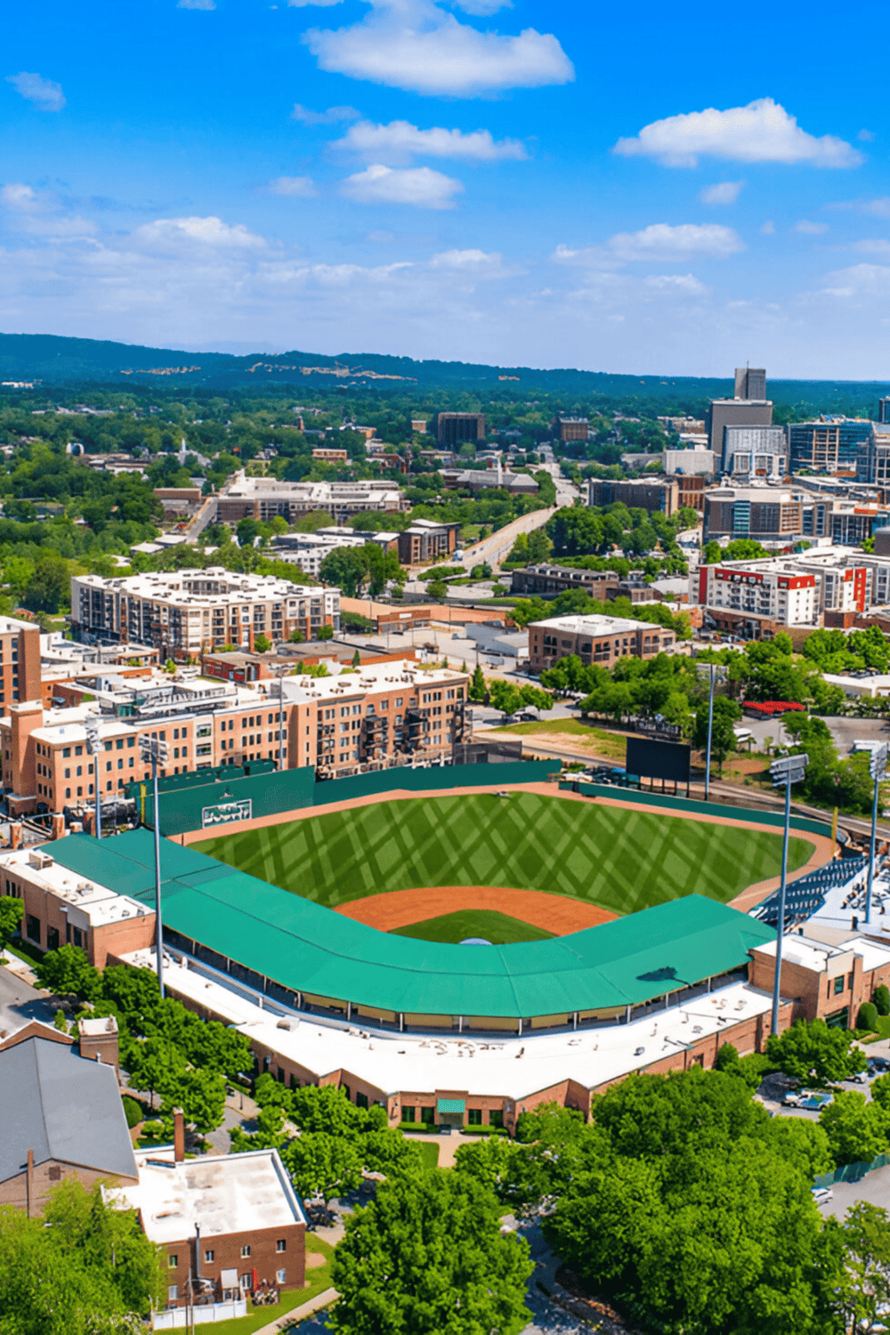 Aerial view of a city with a baseball stadium, skyscrapers, and green parks under a bright blue sky.
