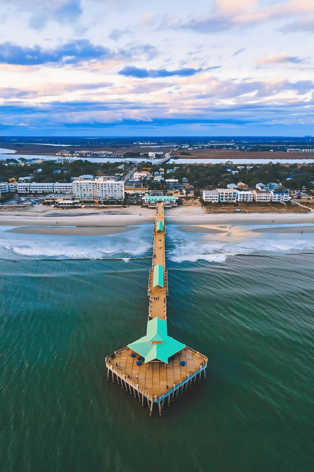 Aerial view of a coastal pier leading to a beach with ocean waves and nearby modern buildings.