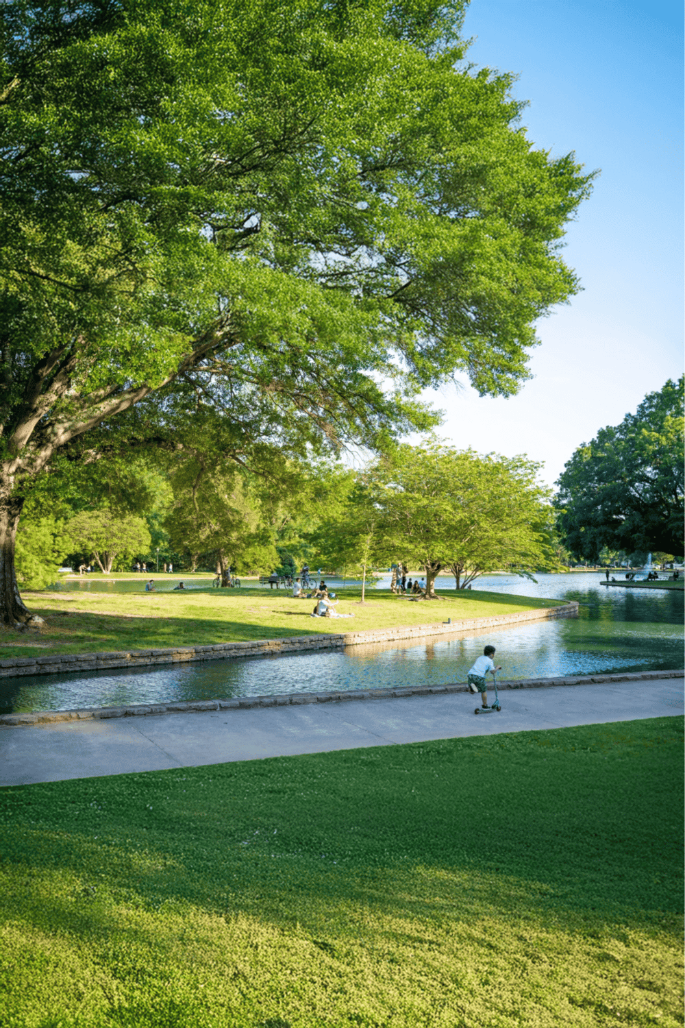 Lush green park with children playing near water, trees, and sunny sky.