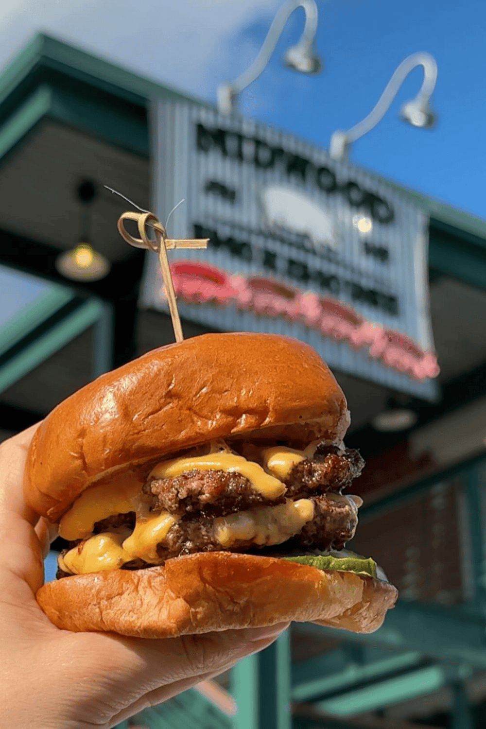 Cheeseburger with multiple beef patties and melted cheese in front of a restaurant sign.