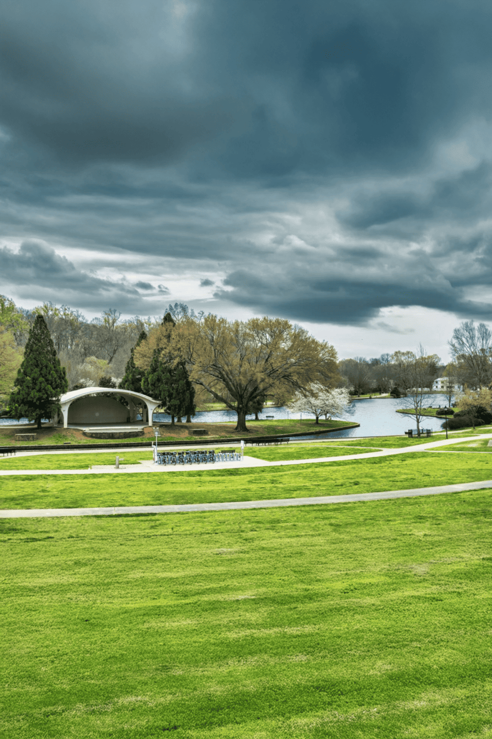 Vibrant park with lush green lawns, trees, and a serene lake under dramatic cloudy sky.