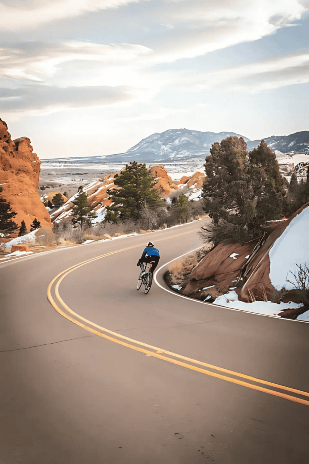 Cyclist riding on scenic mountain road with snow, rocks, and trees at sunset.