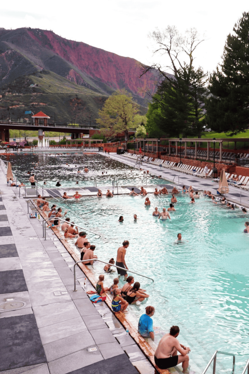 Outdoor hot springs pool with mountain views, surrounded by lush greenery and visitors enjoying the water.