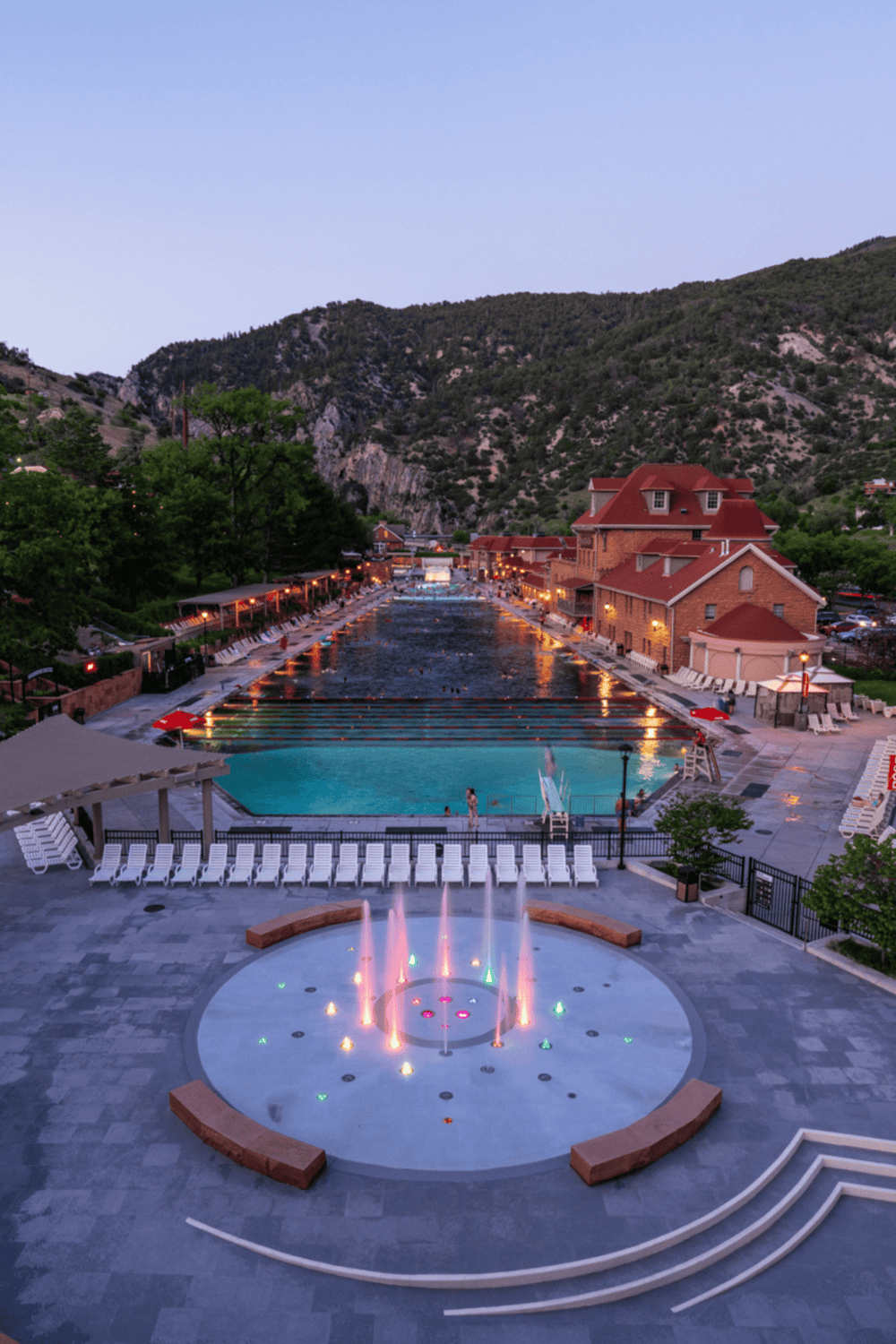 Vibrant resort pool area at dusk surrounded by mountains, with illuminated fountains and outdoor lounge chairs.